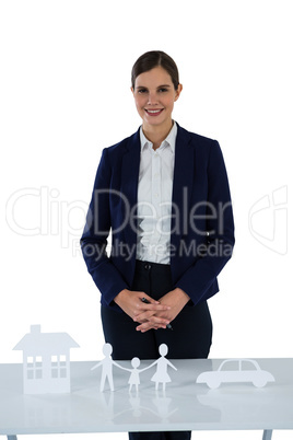 Smiling businesswoman standing with paper cut out of family, house and car on table