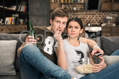 portrait of concentrated couple watching movie with beer and popcorn at home