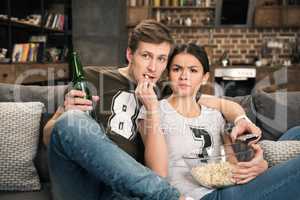 portrait of concentrated couple watching movie with beer and popcorn at home