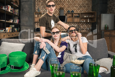 young friends in 3D glasses watching movie on sofa at st patricks day