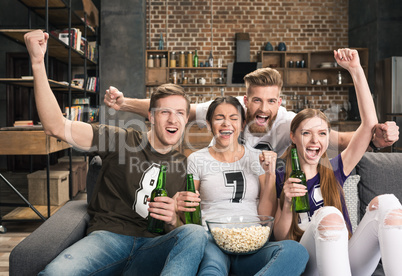 Excited young friends drinking beer and eating popcorn while supporting favorite team at home