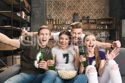 Excited young friends drinking beer and eating popcorn cheering at camera