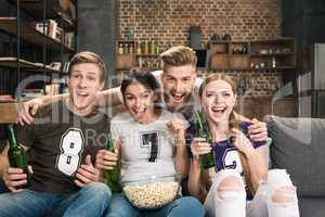 Excited young friends in t-shirts drinking beer and eating popcorn at home