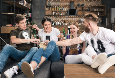 Cheerful young friends drinking beer and eating popcorn on sofa