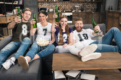 Happy young friends toasting with beer bottles while sitting on sofa and smiling at camera