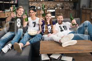 Happy young friends toasting with beer bottles while sitting on sofa and smiling at camera