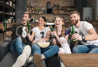 Cheerful young friends holding beer bottles and looking away at home