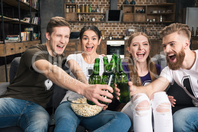 Happy young friends clinking beer bottles while sitting on sofa
