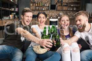 Happy young friends clinking beer bottles while sitting on sofa