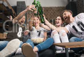 Happy young friends clinking beer bottles while sitting on sofa