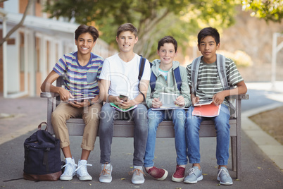 Portrait of school kids using mobile phone and digital tablet on bench in school campus