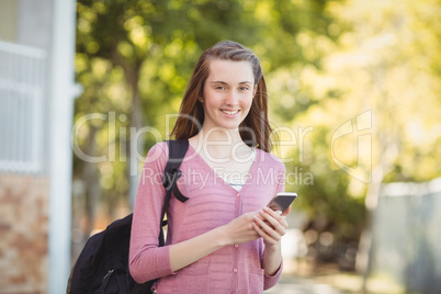 Smiling school girl with schoolbag using mobile phone in campus