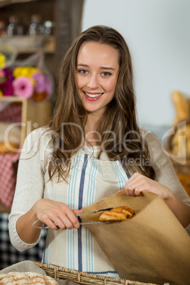 Portrait of smiling female staff putting croissant into a paper bag at counter