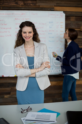 Smiling business executive standing with arms crossed while colleague working in background