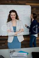 Smiling business executive standing with arms crossed while colleague working in background