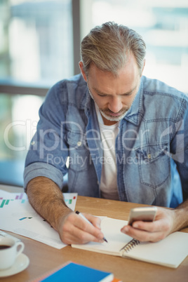 Male executive writing in organizer while using mobile phone Male executive writing in organizer while using mobile phone