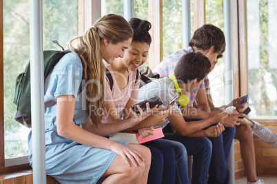 Happy students sitting on window sill and using mobile phone in corridor