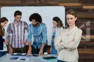 Portrait of businesswoman standing with arms crossed while colleague working in background