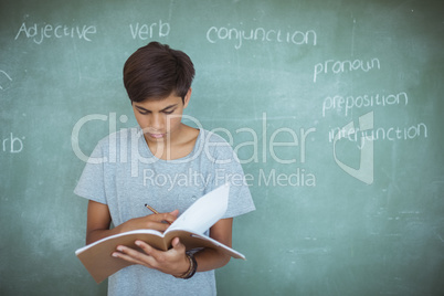 Attentive schoolboy reading book against chalkboard in classroom