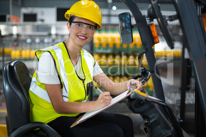 Female factory worker sitting on forklift and writing in clipboard at factory