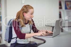Happy schoolgirl with schoolbag using laptop in library