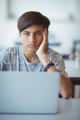 Portrait of serious schoolboy sitting with laptop in classroom