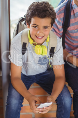 Portrait of happy schoolboy sitting on window sill and using mobile phone in corridor