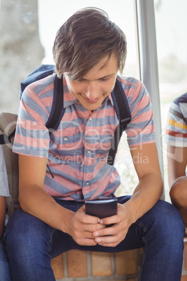 Happy schoolboy sitting on window sill and using mobile phone in corridor