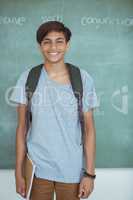 Smiling schoolboy with backpack and book standing against chalkboard in classroom