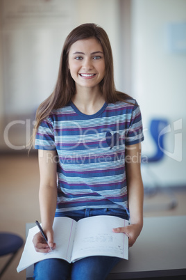 Portrait of schoolgirl sitting on bench and writing on book in classroom