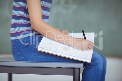 Mid section of schoolgirl sitting on bench and writing on book in classroom