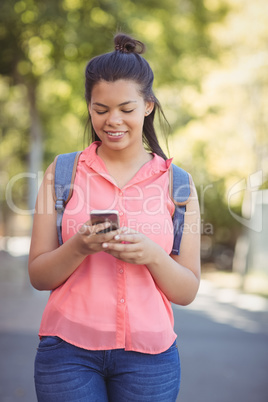 Smiling school girl with schoolbag using mobile phone in campus