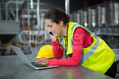 Female factory worker using laptop while talking on mobile phone