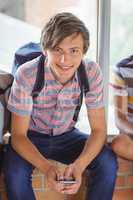 Portrait of happy schoolboy sitting on window sill and using mobile phone in corridor