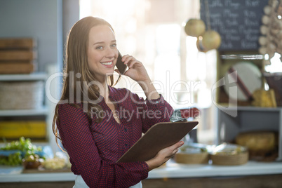 Smiling female staff holding clipboard and talking on mobile phone at counter