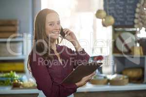 Smiling female staff holding clipboard and talking on mobile phone at counter