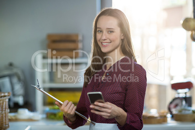 Portrait of smiling female staff holding clipboard and using mobile phone at counter