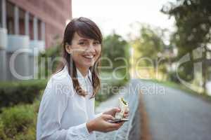 Smiling executive having veg roll while using mobile phone on road