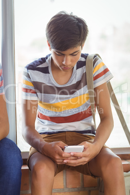 Attentive schoolboy sitting on window sill and using mobile phone in corridor