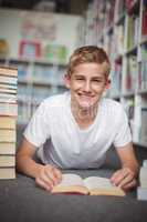 Portrait of happy schoolboy lying on floor with books in library