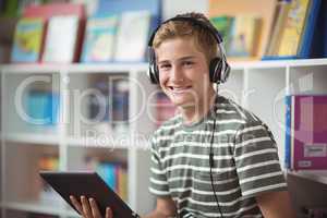 Portrait of happy schoolboy listening music while using digital tablet in library