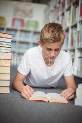 Attentive schoolboy lying on floor and reading book in library