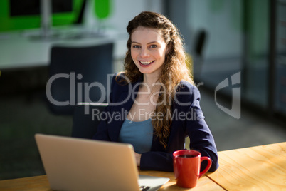 Smiling female business executive sitting at desk with laptop and coffee cup