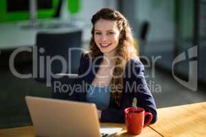 Smiling female business executive sitting at desk with laptop and coffee cup