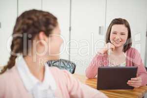 Smiling schoolgirl with digital tablet talking to her friend in classroom