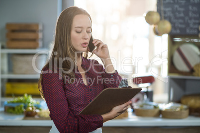 Female staff checking clipboard while talking on mobile phone at counter