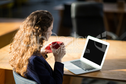 Female business executive looking at laptop while having cup of coffee