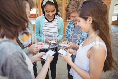 Smiling school students standing in circle and using mobile phone