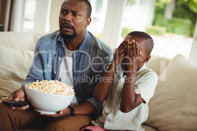 Son covering his eyes while watching television with father