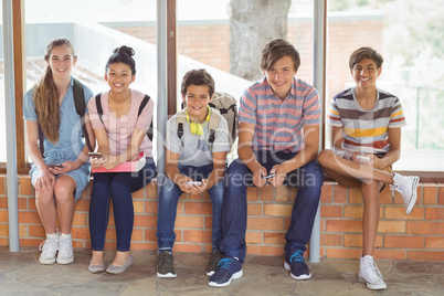Portrait of happy students sitting on window sill and using mobile phone in corridor